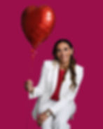 A smiling woman in a white suit and red top holds a red heart-shaped balloon against a solid magenta background.