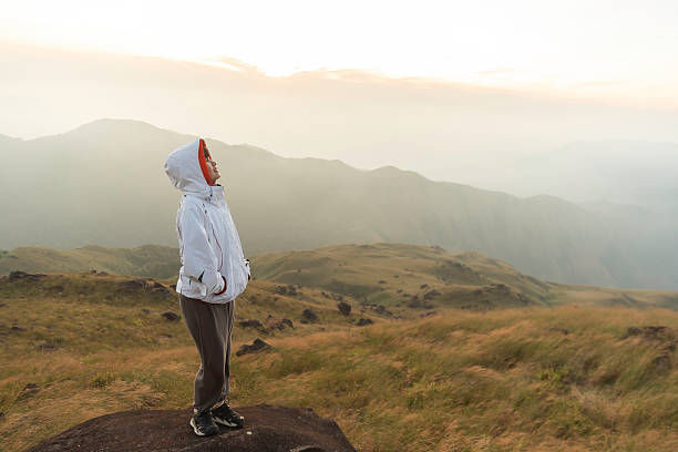 Person in a white jacket stands on a rock, gazing at misty mountains during sunrise. The scene is serene with muted earth tones.