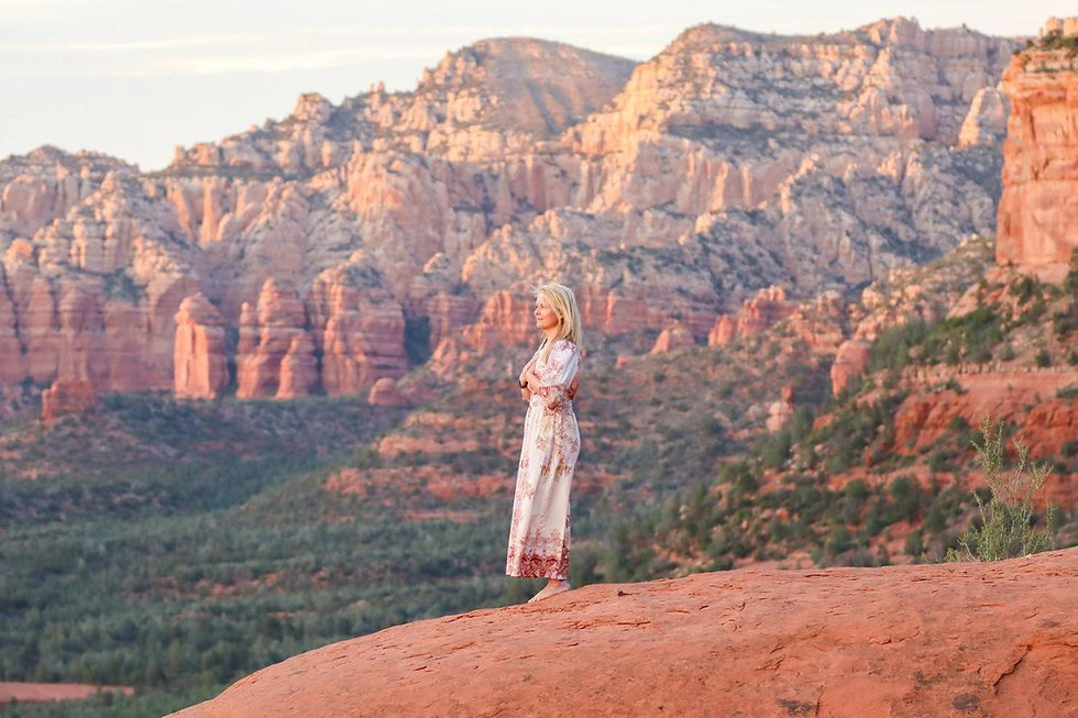 Blonde woman in a floral dress stands on a red rock ledge before sunlit canyon cliffs at sunset, calm and reflective.