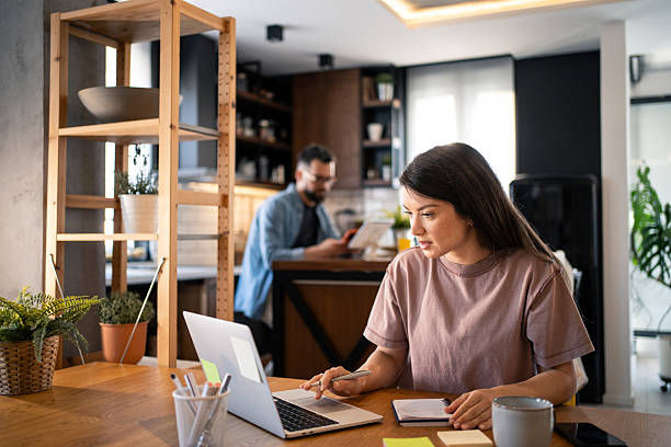 Woman working on a laptop at a wooden table in a cozy kitchen, with a man reading in the background. Bright and focused atmosphere.