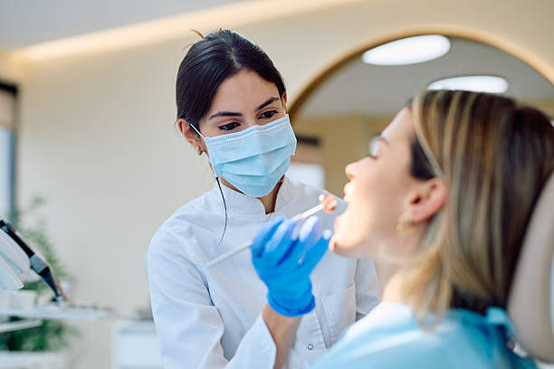 Masked dentist examines a woman’s mouth with a tool in a bright clinic, focused and calm during a dental checkup