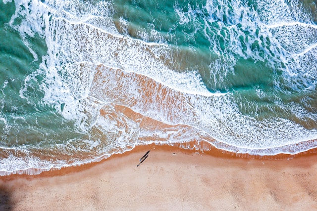 Aerial view of a sandy beach with foamy waves. Two people walk along the shore, casting long shadows. Turquoise sea contrasts with tan sand.
