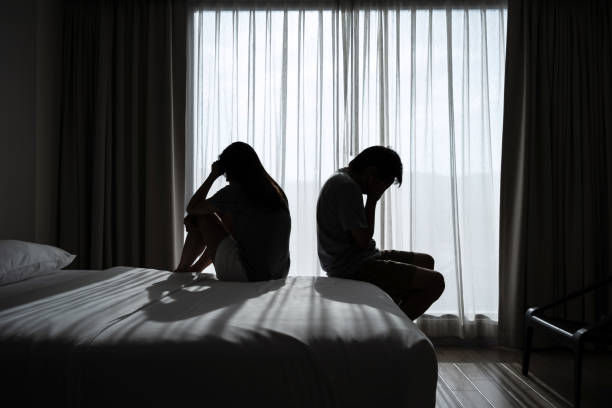 Silhouetted couple sits back-to-back on a bed, appearing upset. Sunlight filters through curtains in a dimly lit room.
