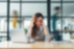 Stressed woman at a laptop in a glass-walled office, hands on temples, with blurred coworkers moving behind her.