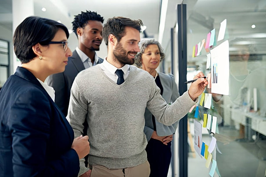 Four people in an office brainstorm with colorful sticky notes on a glass wall. A man draws on a chart, creating a collaborative mood.