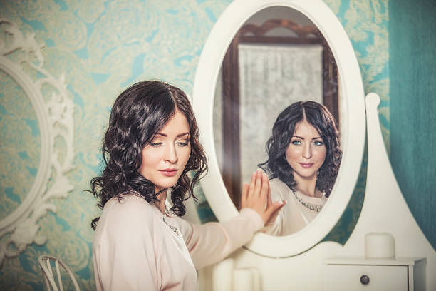 Woman touches oval mirror, sees reflection smiling. Vintage setting with ornate wallpaper. Calm, introspective mood.