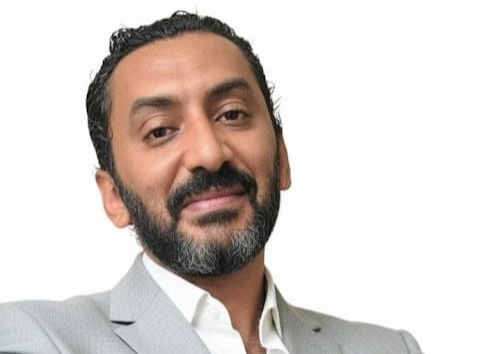 Man with a beard, wearing a light gray suit and white shirt, smiles with a confident expression against a plain white background.