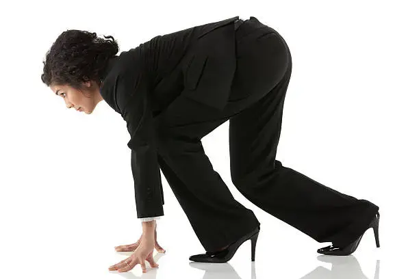 A woman in a black suit crouches in a starting position, poised to run. She stands on a reflective white surface, suggesting readiness.