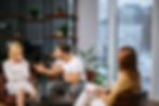 A woman cries while a man gestures during a counseling session. Another woman observes, writing notes. Shelves and plants in the background.