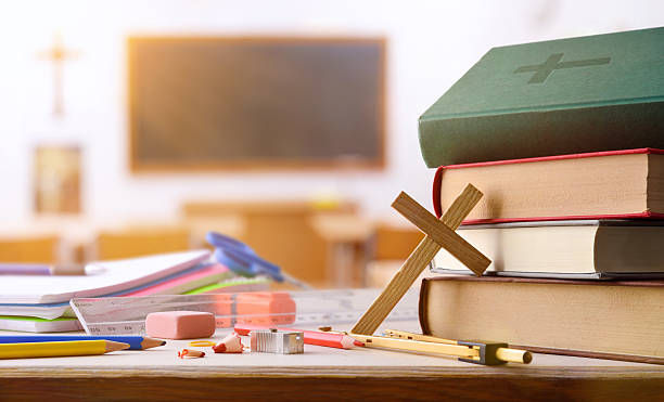 Stack of religious books with a wooden cross on a desk, surrounded by school supplies. Background features a blurred classroom with a chalkboard.