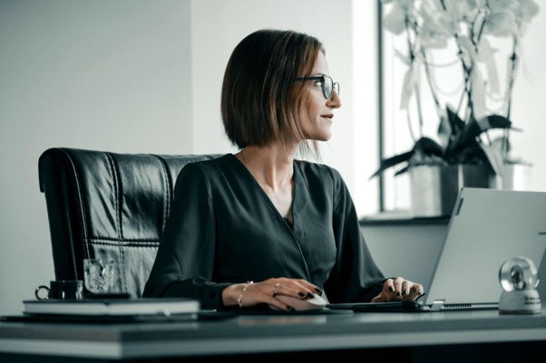 Woman in glasses and black blouse sits at an office desk using a computer, looking right in a calm modern workspace.