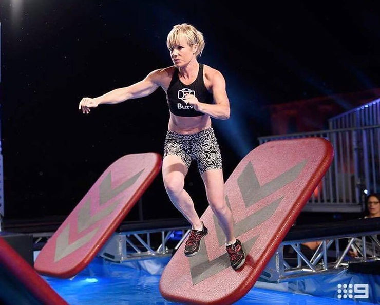 Athlete jumps between tilted boards in a competition, wearing a black top and patterned shorts. Focused expression, colorful lights around.