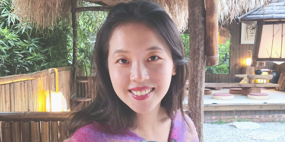 Smiling woman wearing a purple top in a cozy outdoor setting with bamboo, greenery, and soft lighting, creating a warm ambiance.