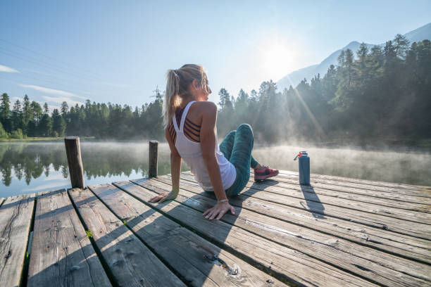 Woman in activewear relaxes on a lakeside dock, facing misty forest and mountains at sunrise. Water bottle beside her. Peaceful morning.