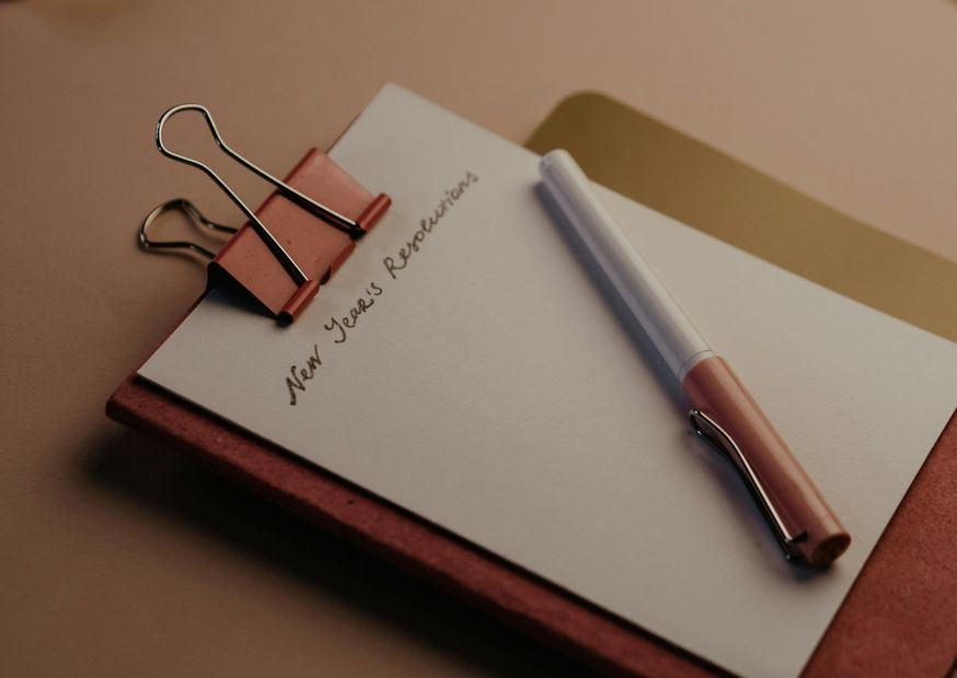 Clipboard with "New Year's Resolutions" written on paper, held by a red clip. A pen rests beside it on a brown surface. Cozy atmosphere.