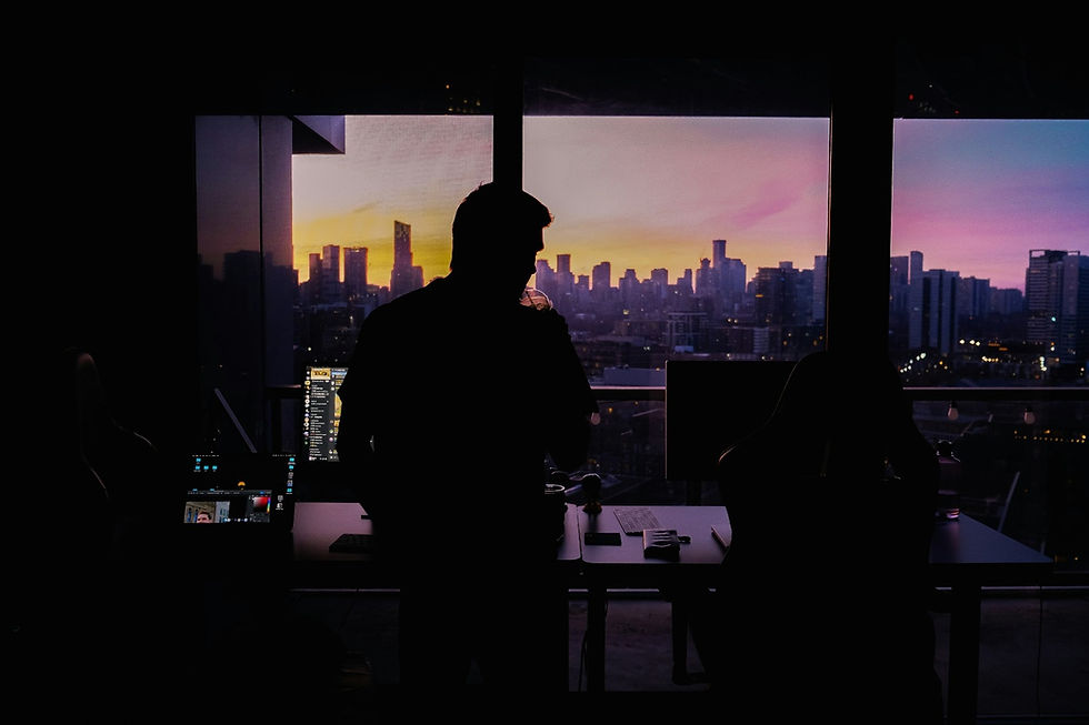 Silhouette of a person in a dark room with cityscape view at sunset. Bright screens and a purple-orange sky set a contemplative mood.
