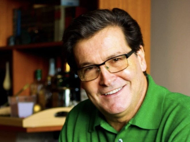 Smiling man with glasses wearing a green shirt, sitting indoors. Background has shelves with books and bottles, creating a relaxed mood.