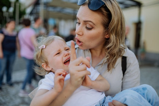 Woman calming crying child outdoors, gesturing gently. The background shows blurred people, creating a lively atmosphere.
