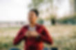 Person in a red jacket sitting outdoors with hands on chest, eyes closed, meditating. Grass and trees in background. Calm mood.