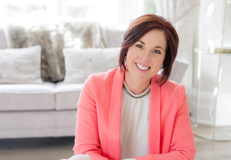 Smiling woman in a coral blazer sits in a bright room with a beige sofa and cushions in the background. The mood is cheerful and welcoming.