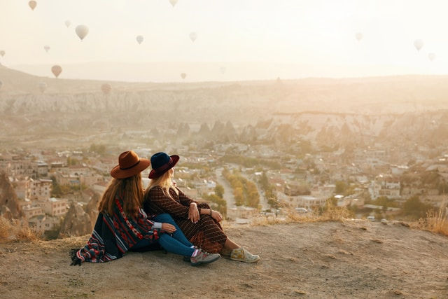 Two people in hats sit on a cliff overlooking a cityscape at sunset, with hot air balloons in the sky, creating a serene atmosphere.