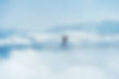 A person walks on a high rope bridge above clouds, with mountains in the background. It's a bright, clear day, evoking a sense of adventure.