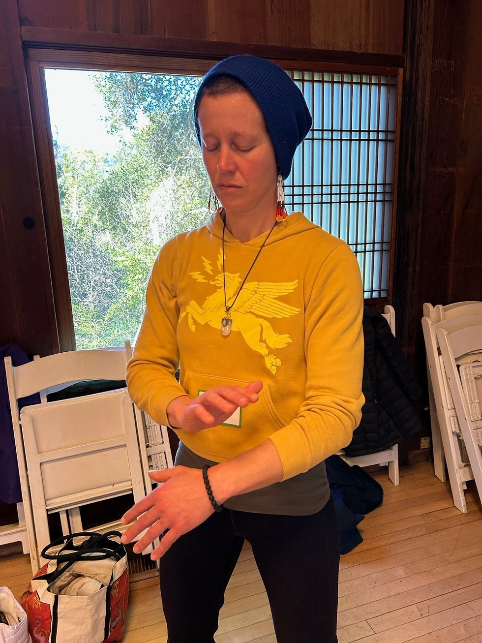 Woman in a yellow hoodie and blue beanie stands with eyes closed, meditating in a wooden room with chairs and a bright window.