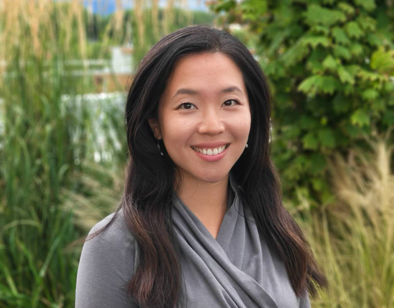 Smiling woman in a gray top stands outdoors among green plants and tall grasses.