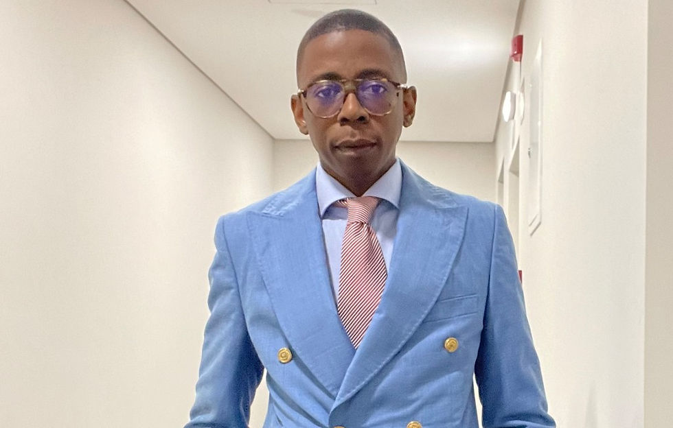 Man in a light blue suit and pink striped tie stands in a bright hallway, wearing glasses and a serious expression.