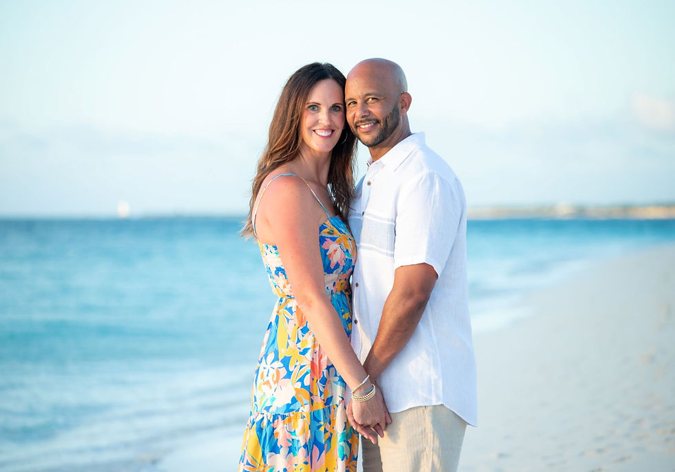 Couple holding hands and smiling on a beach. Woman in floral dress, man in white shirt. Ocean and sky in the background.