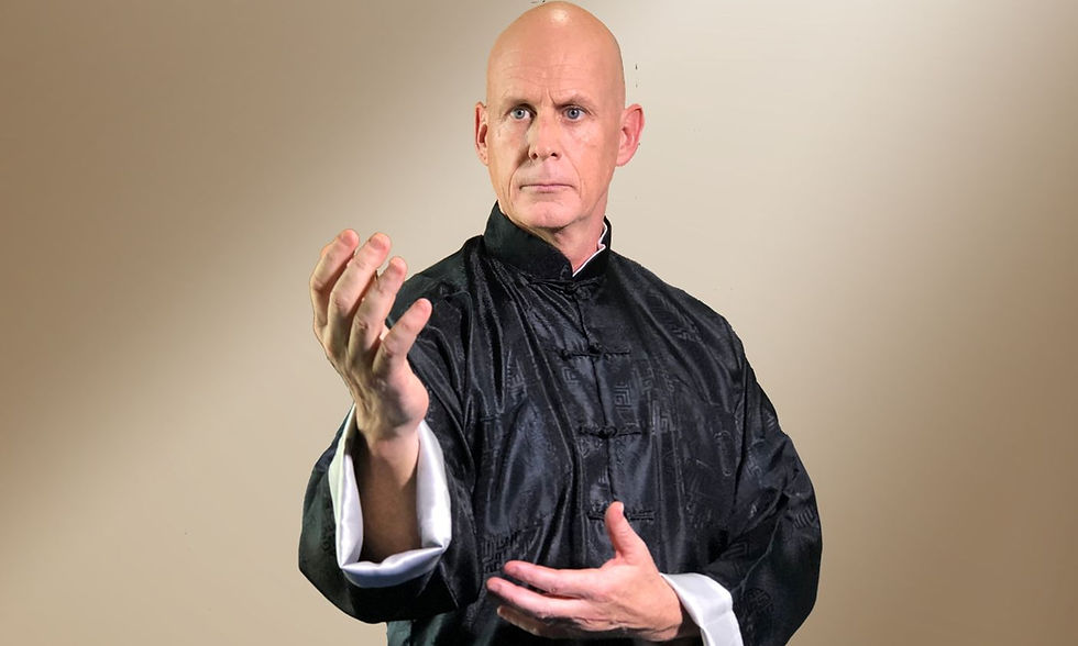 Bald man in traditional black martial arts attire demonstrates a hand pose against a neutral background, exuding focus and calm.