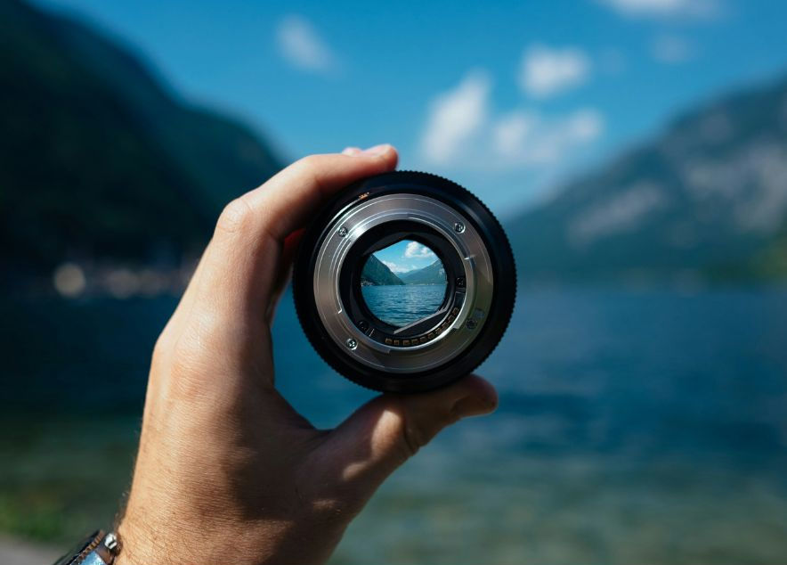 Hand holds camera lens with lake and mountains in focus through the lens. Blue sky and water in the blurred background. Peaceful scene.