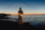Silhouetted person on a beach at dusk, surrounded by glowing spiral light trails over calm water.