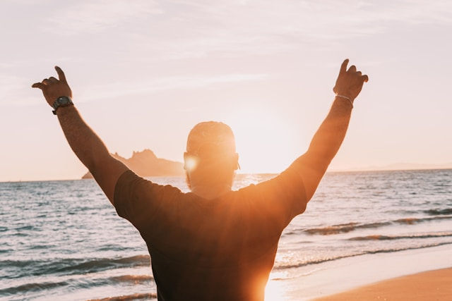 Person seen from behind on a beach at sunset, arms raised in celebration as waves roll in and warm light glows.