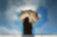 A person holding an umbrella is reflected in a puddle against a bright blue sky with clouds.