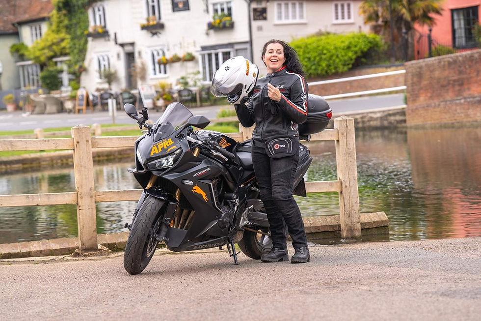 Woman in black gear holding helmet, smiling by a black motorcycle with "APRIL" text. Scenic village background with a pond and greenery.