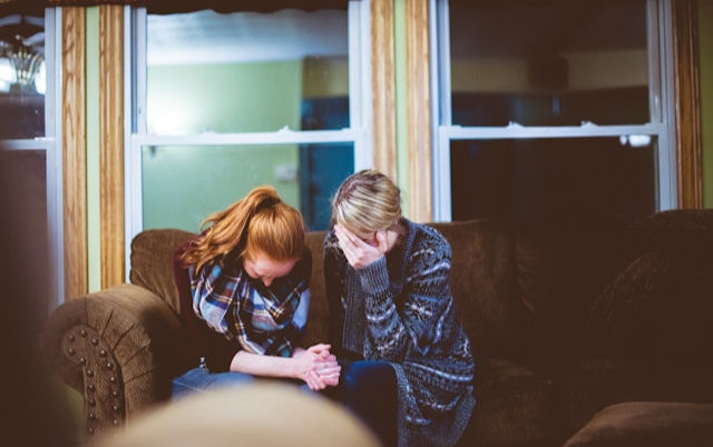 two women sitting on a couch, both appearing emotionally distressed, with one covering her face and the other leaning forward