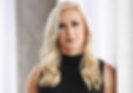 Blonde woman in a black top with wavy hair stands against a light curtain. She has a neutral expression. Background is concrete and white.