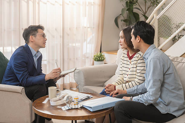 A man consults a seated couple in a living room. Papers, a pen, and a coffee cup are on a table. Neutral tones, relaxed atmosphere.