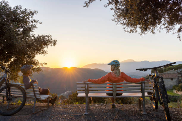Two cyclists wearing helmets sit on benches, facing a scenic sunset over mountains. Bicycles are nearby, trees frame the view, creating a peaceful mood.