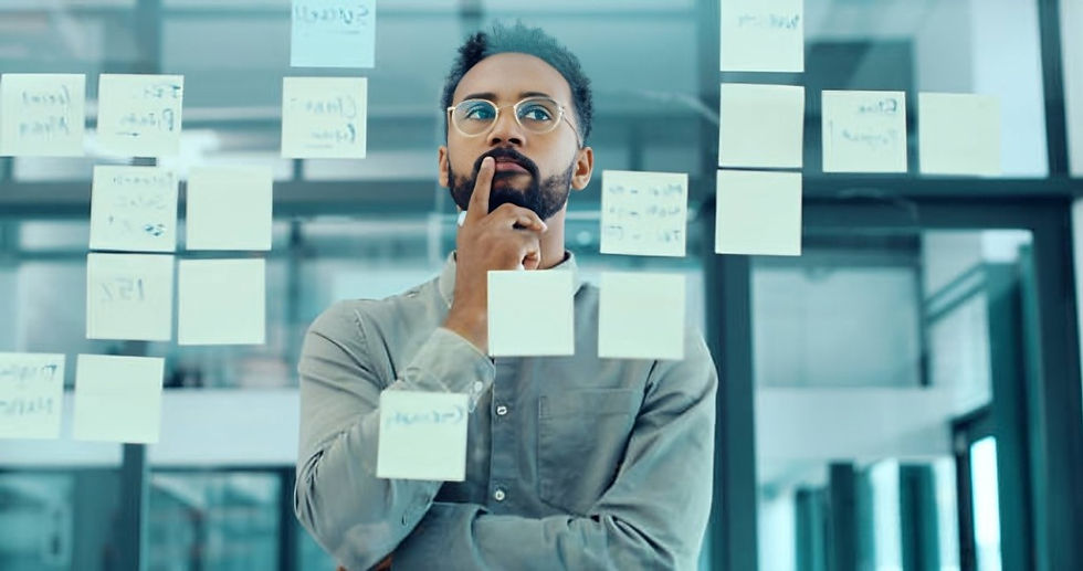 Man contemplating in front of glass wall with sticky notes in office. He's wearing glasses and a gray shirt, conveying a thoughtful mood.