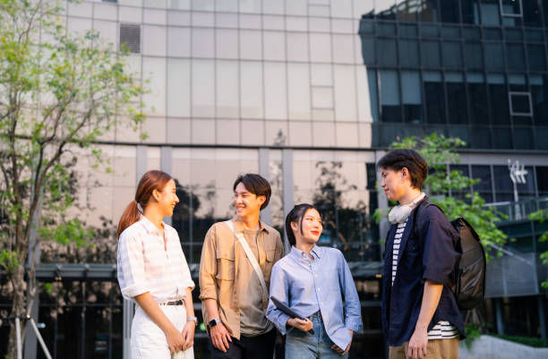 Four people smiling and chatting outside a glass building. Trees nearby; casual attire evokes a relaxed, friendly mood.