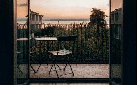 A serene sunset view from a balcony with a table and chair. Overlooks a lush garden and distant water, with warm orange and pink hues.