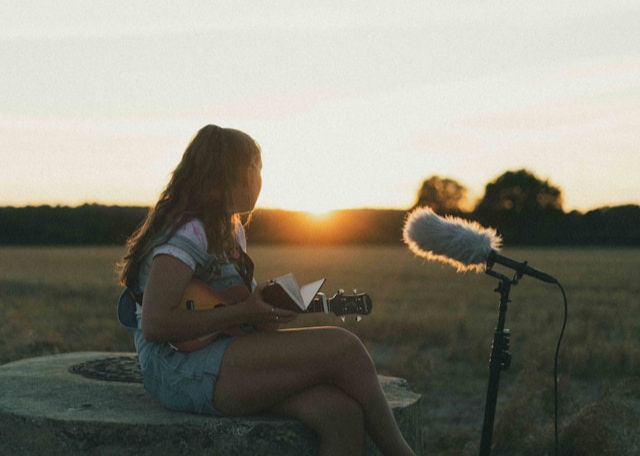 A young woman sits on a stone, playing a ukulele and singing into a microphone as the sun sets in a field.