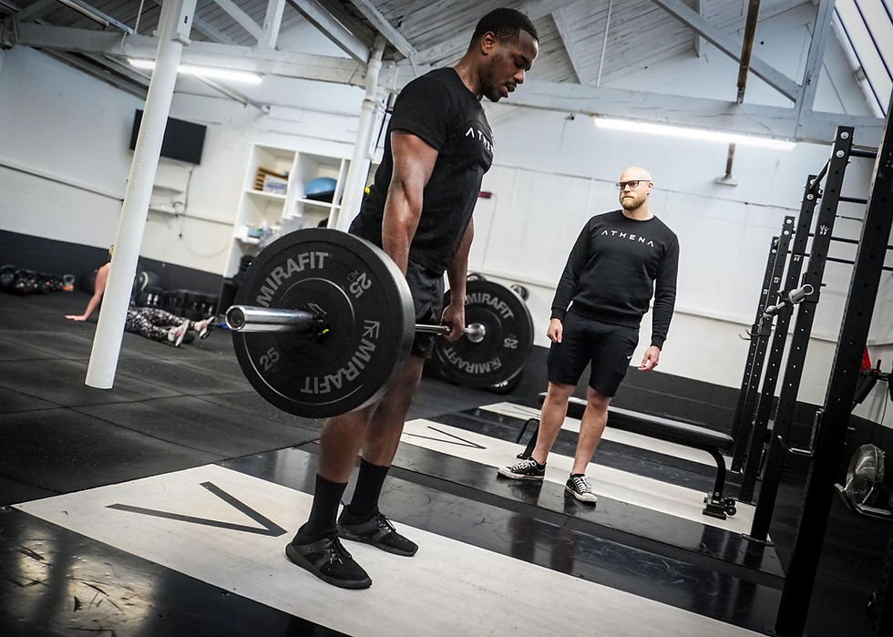 Black-clad man deadlifts a barbell in a gym while a coach watches; ATHENA and MIRAFIT text visible.