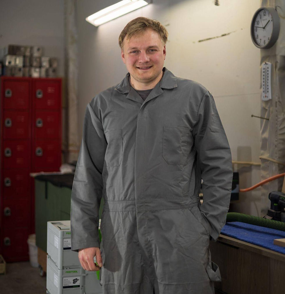 Smiling person in gray coverall standing in a workshop. Red lockers, a clock, and green equipment boxes visible in the background.