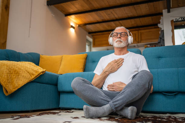 Elderly man sitting cross-legged, eyes closed, with headphones on. He’s meditating with hands on chest and belly, next to a blue sofa with yellow cushions. Cozy room.