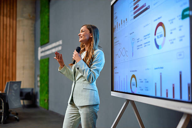 A woman in a light suit holds a microphone, presenting data charts on a screen in a modern conference room, conveying confidence and focus.