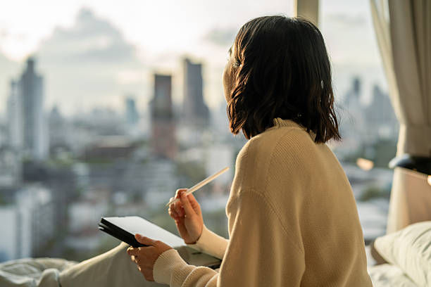 Person in a sweater sits on a bed, holding a tablet and stylus, gazing out a window at a blurred cityscape at sunset. Cozy and contemplative mood.