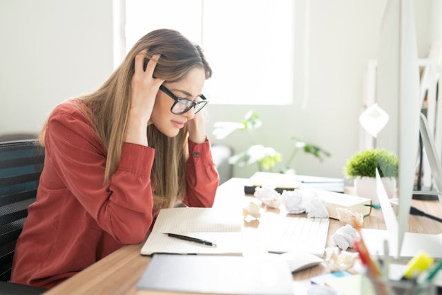 Woman in red shirt, wearing glasses, looks stressed at desk with crumpled papers and notebook. Bright room with plants in background.